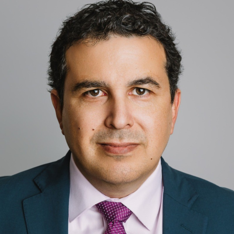 Professional headshot of man in dark suit and purple tie against gray background